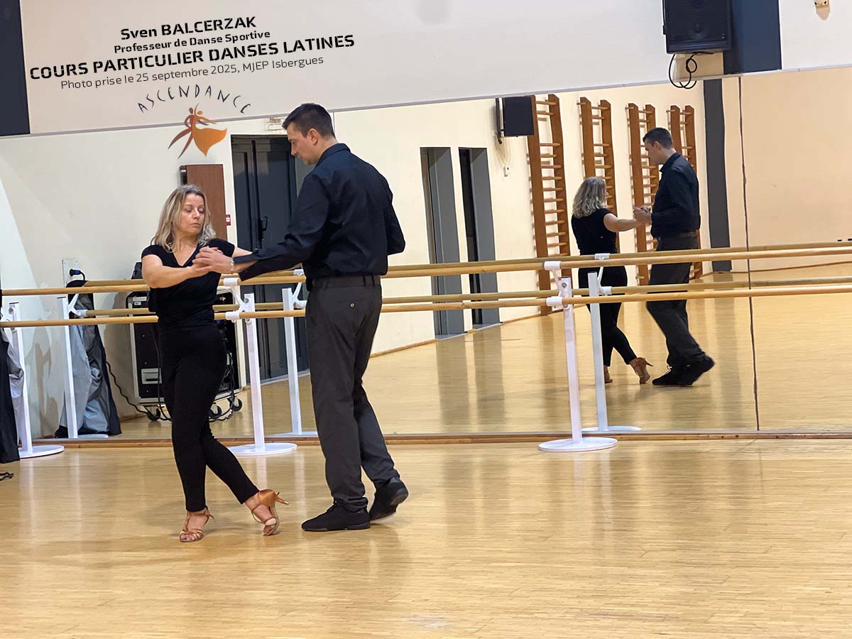 Sven BALCERZAK corrige Virginie dans une figure de Samba à la MJEP DANSE d’Isbergues, sur parquet en bois avec miroirs muraux.