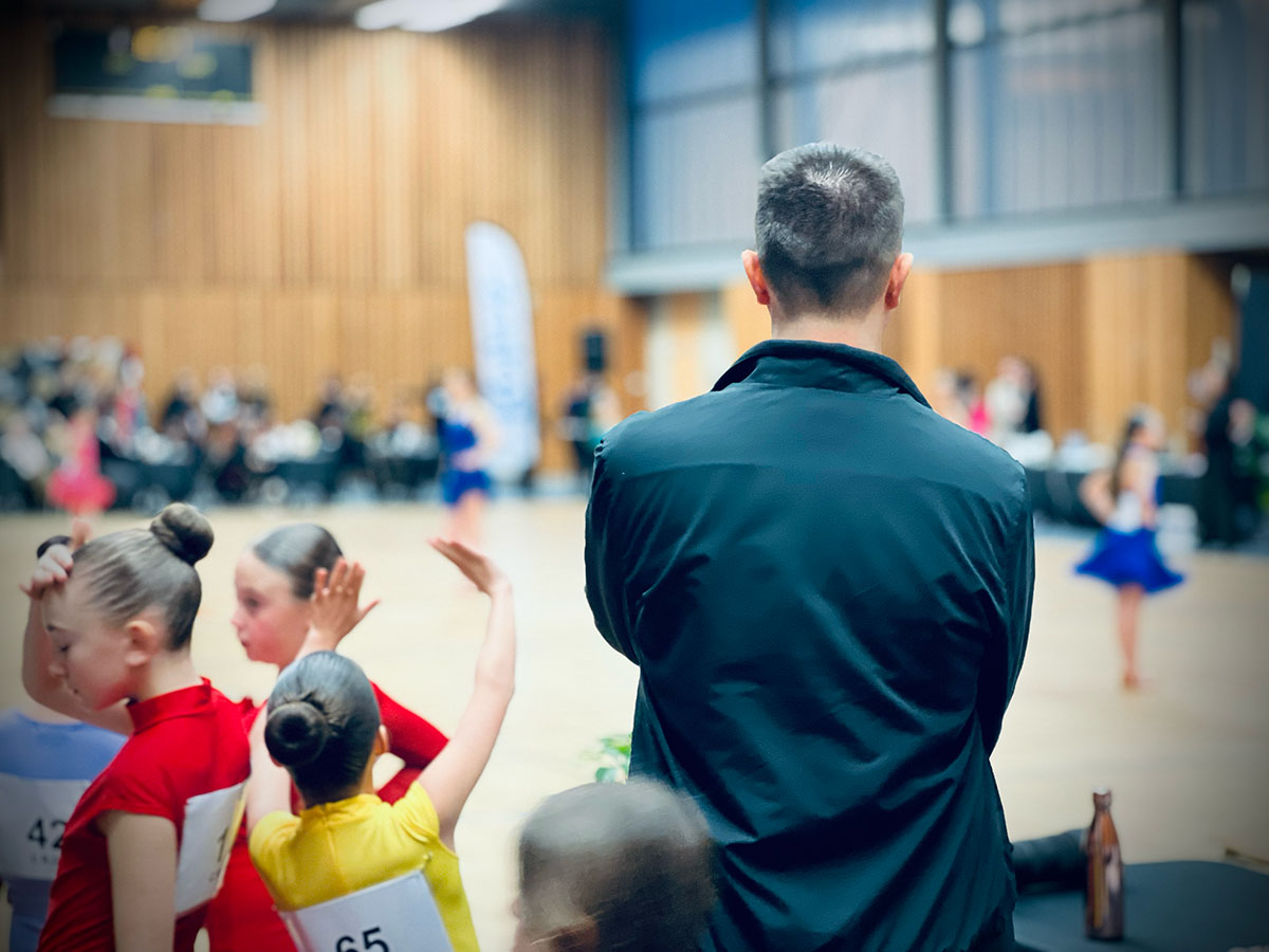Sven Balcerzak de dos, observant ses compétiteurs d’ASCENDANCE sur la piste de danse au Trophée des Sacres de Danse Sportive à Reims, prêt à leur donner un feedback et les coacher pour le prochain tour.